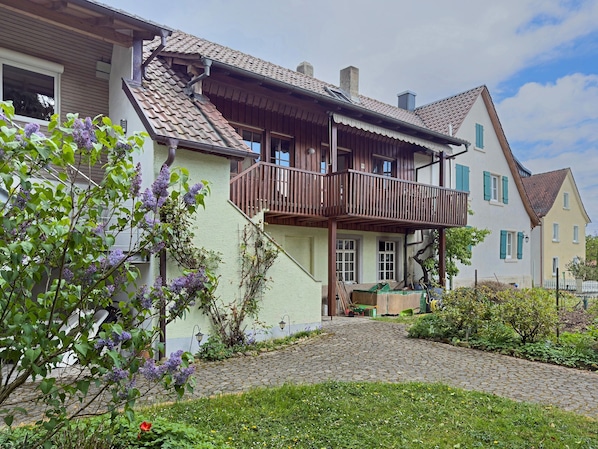 A traditional European house with a wooden balcony, a cobblestone driveway, and a garden with flowering plants.