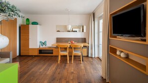 A modern living room with a wooden dining table, two chairs, a TV mounted on a wall, and a large window. - Obst- und Ferienhof Lehle – Apartment Jonagold with Shared Terrace, Garden & Wi-Fi (Immenstaad am Bodensee)