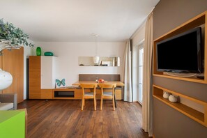 A modern living room with a wooden dining table, two chairs, a TV mounted on a wall, and a large window.