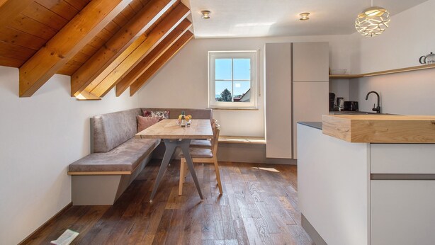 A loft-style room with a wooden ceiling, a dining table, a bench, and a kitchen area with a window.