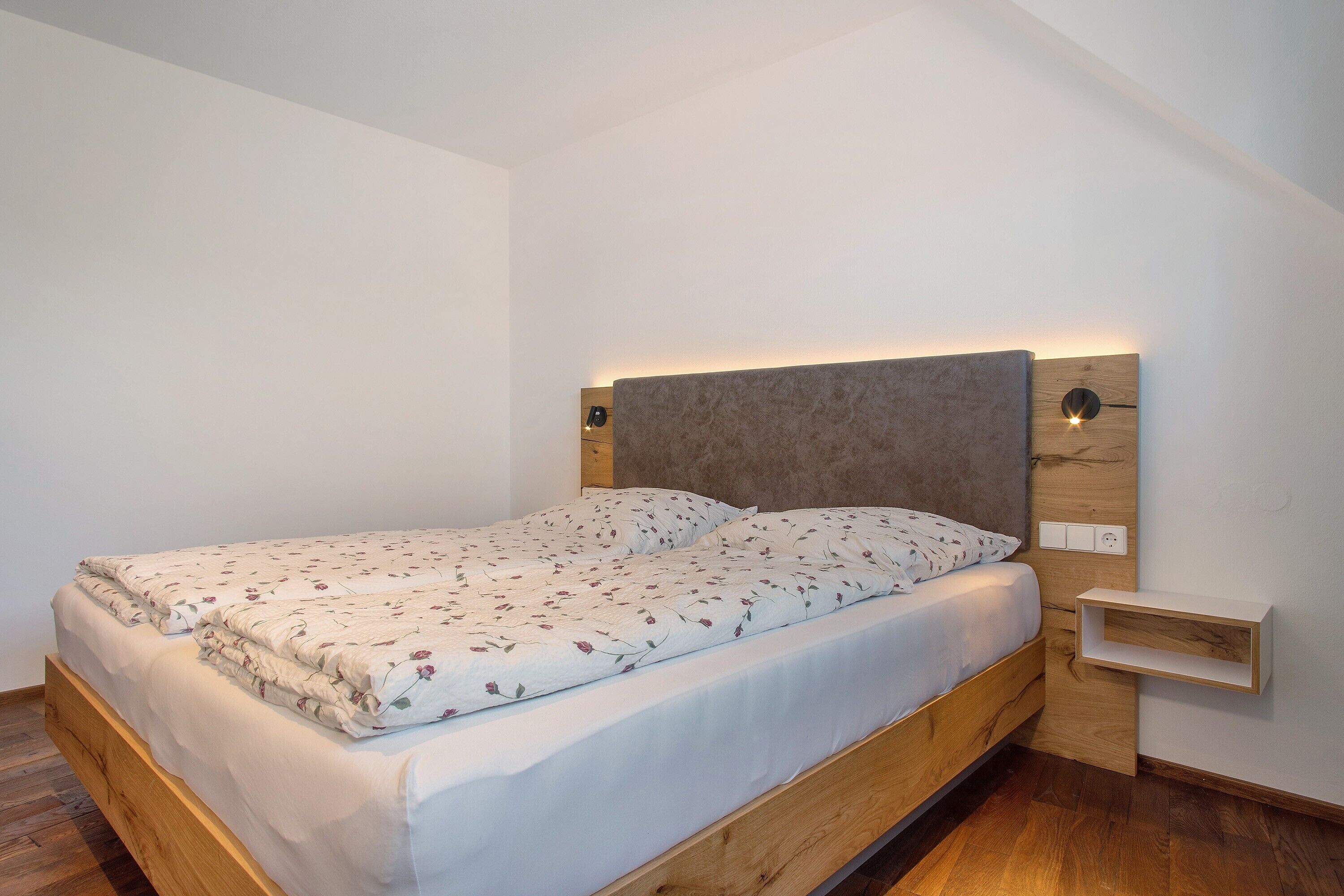A single bed with a wooden frame, a headboard with integrated lighting, a bedside table, and a small shelf.