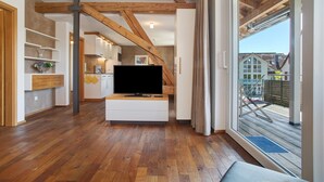 Ein modernes Wohnzimmer mit Flachbildfernseher, Holzbalken, einer Schiebeglastür zum Außenbereich und einer Küche im Hintergrund.