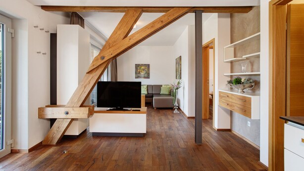 Ein modernes Interieur mit Holzbalkendecke, einem Flachbildfernseher auf einer weißen Konsole, einer Couch und einer Küchenzeile mit Holzschränken und einer Dekorecke.