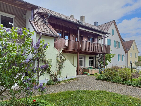 Une maison européenne traditionnelle avec un balcon en bois, une allée pavée et un jardin agrémenté de plantes à fleurs.