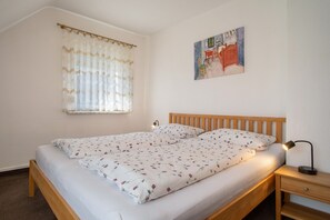 Un lit simple avec une tête de lit en bois, une table de chevet avec une lampe, une fenêtre avec des rideaux légers et un tableau accroché au mur.