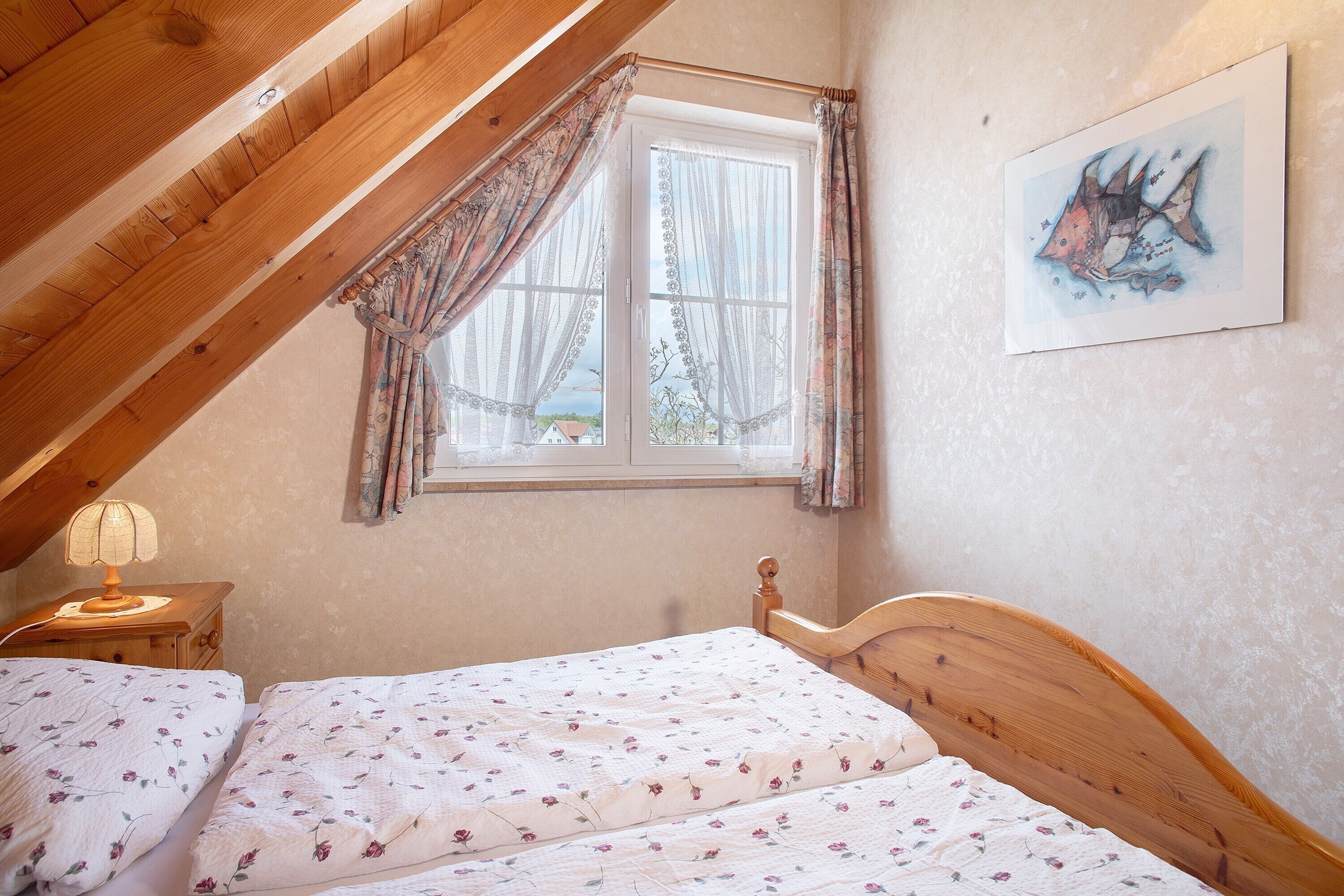 Une chambre avec un plafond en bois, un lit recouvert d’une literie à motifs floraux, une table de chevet avec une lampe et une fenêtre avec des rideaux.