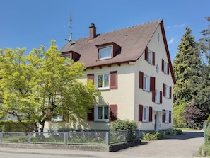 Une maison à deux étages, dotée d’un toit en pente, de nombreuses fenêtres avec volets et d’une cheminée bien visible.