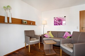 A living room with a sofa, two armchairs, a small table, a floor lamp, a wall-mounted shelf with vases, and a large floral painting.