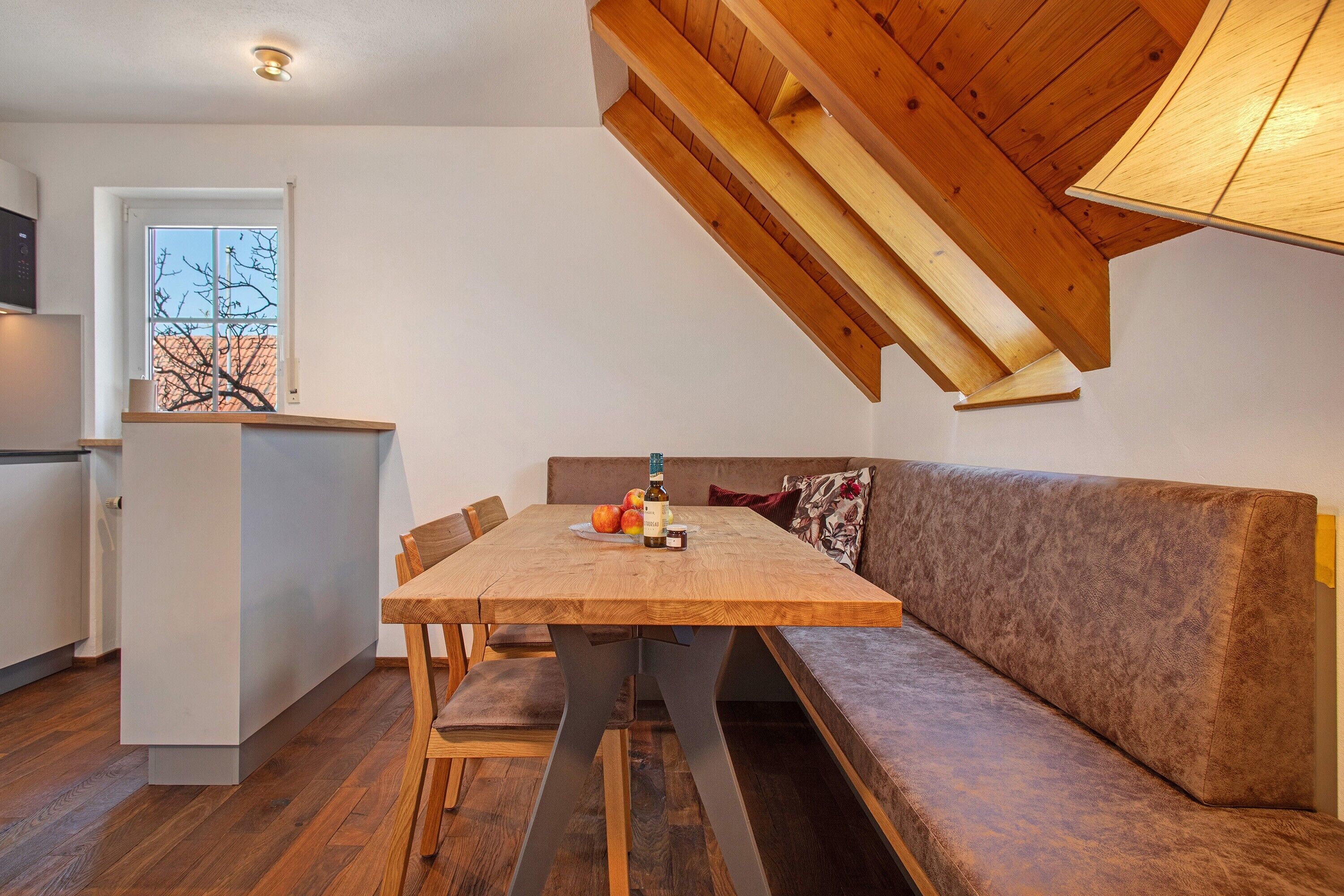A dining area with a wooden table and bench, a kitchen in the background, and a sloped ceiling.