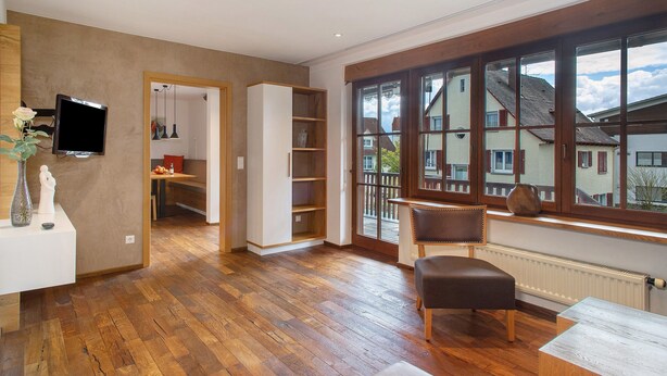 A spacious room with a wooden floor, a television mounted on the wall, a large window with a view of neighboring houses, and a built-in bookshelf. - Obst- und Ferienhof Lehle – Apartment Alte Laube with Shared Terrace, Garden & Wi-Fi (Immenstaad am Bodensee)