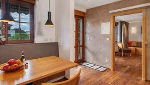 A dining area with a wooden table, chairs, a bowl of fruit, and a bottle. There is a window with a view of houses and a door leading to another room. - Obst- und Ferienhof Lehle – Apartment Alte Laube with Shared Terrace, Garden & Wi-Fi (Immenstaad am Bodensee)
