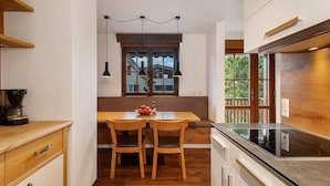 A compact kitchen with a wooden dining table, two chairs, a built-in oven, and a window with a view of greenery. - Obst- und Ferienhof Lehle – Apartment Alte Laube with Shared Terrace, Garden & Wi-Fi (Immenstaad am Bodensee)