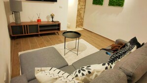A modern living room with a grey sofa, a small black side table, a wooden entertainment unit, a flat-screen TV, and two green wall decorations. - San Giovanni Suite Central (Cagliari)