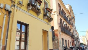 A row of yellow and pink buildings with balconies, a street with parked scooters, and visible electrical boxes. - San Giovanni Suite Central (Cagliari)