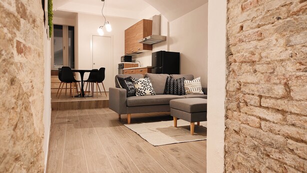 A modern living room with a grey sofa, black and white patterned cushions, a dining area with a table and chairs, and a kitchen in the background. - San Giovanni Suite Central (Cagliari)