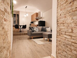 Una sala de estar moderna con un sofá gris, cojines estampados en blanco y negro, una zona de comedor con una mesa y sillas, y una cocina al fondo.
