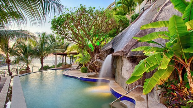A pool with a waterfall feature, surrounded by tropical plants and a stone structure.