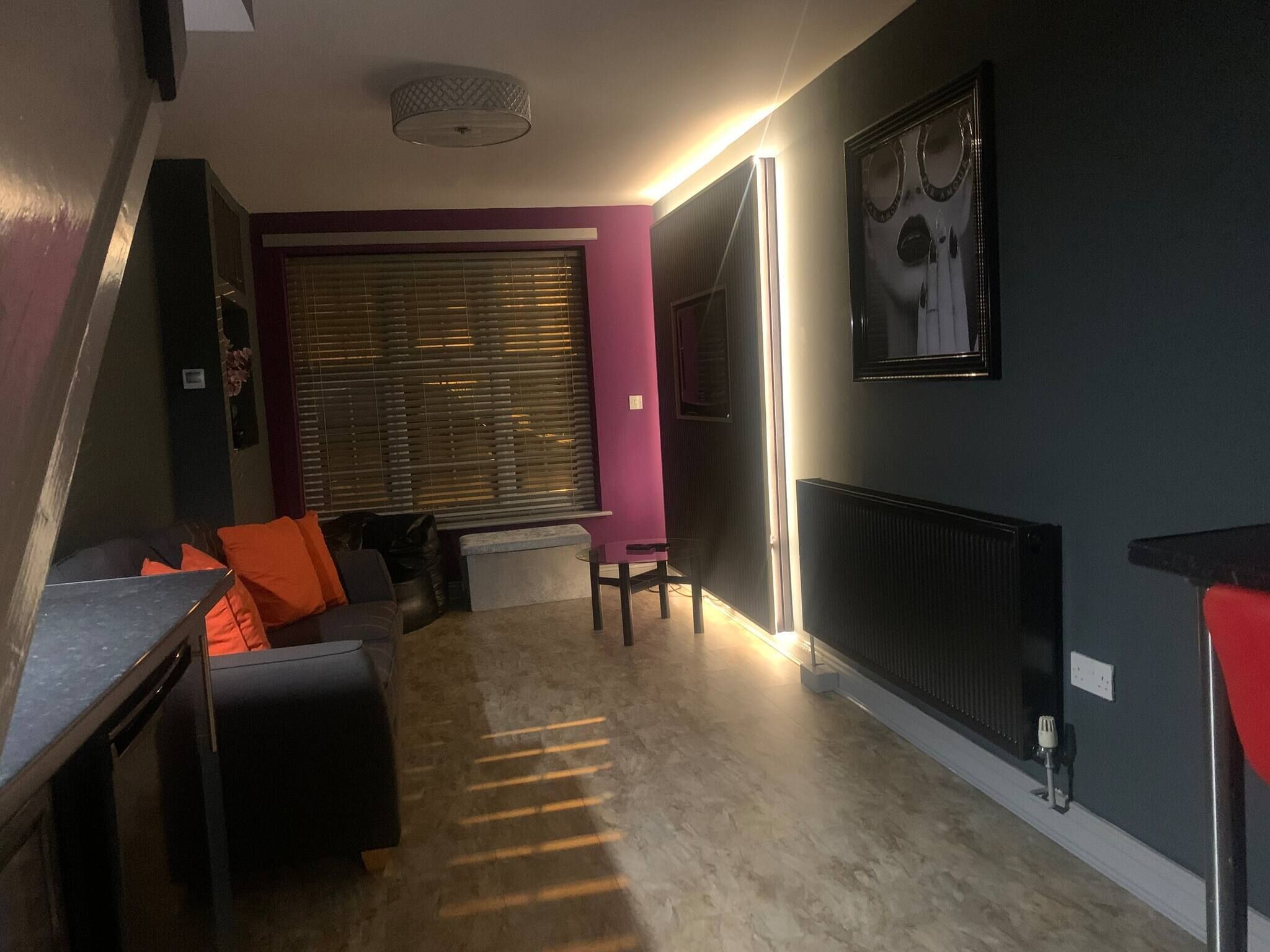 A modern living room with a sofa, ottoman, and a dining table. There is a large window with blinds, a radiator, and a ceiling light fixture.