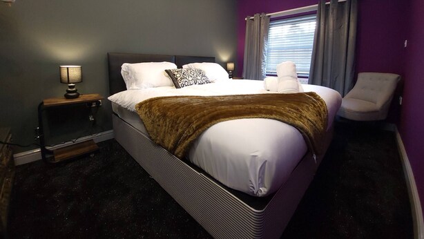 A modern hotel room with a large bed, a bedside table with a lamp, a chair, and a window with curtains. - Liverpool City, Fairfield Duplex Suite (Liverpool)