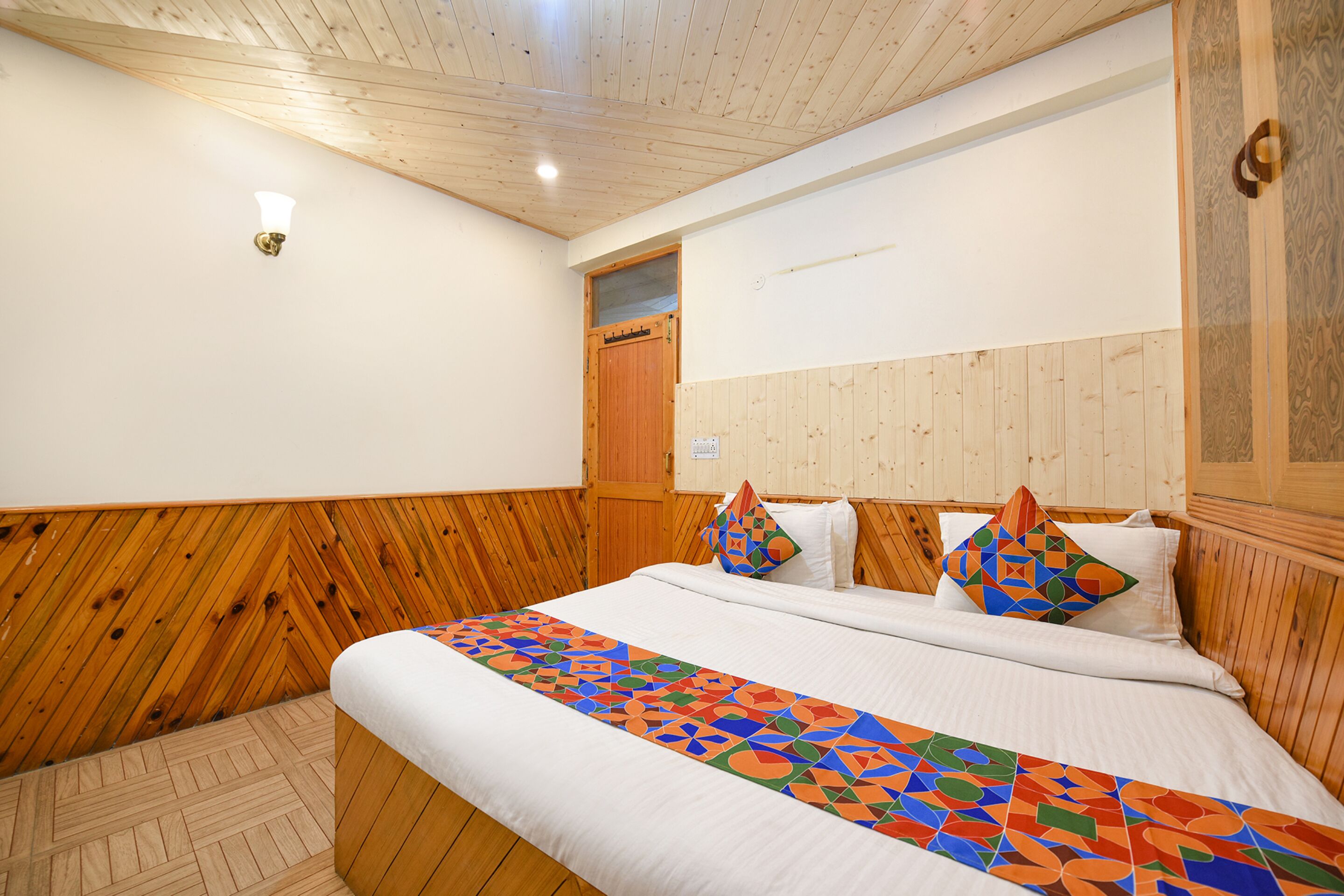 Een slaapkamer met een houten hoofdbord, een bed met wit beddengoed en een kleurrijke, geometrisch gemêleerde deken.