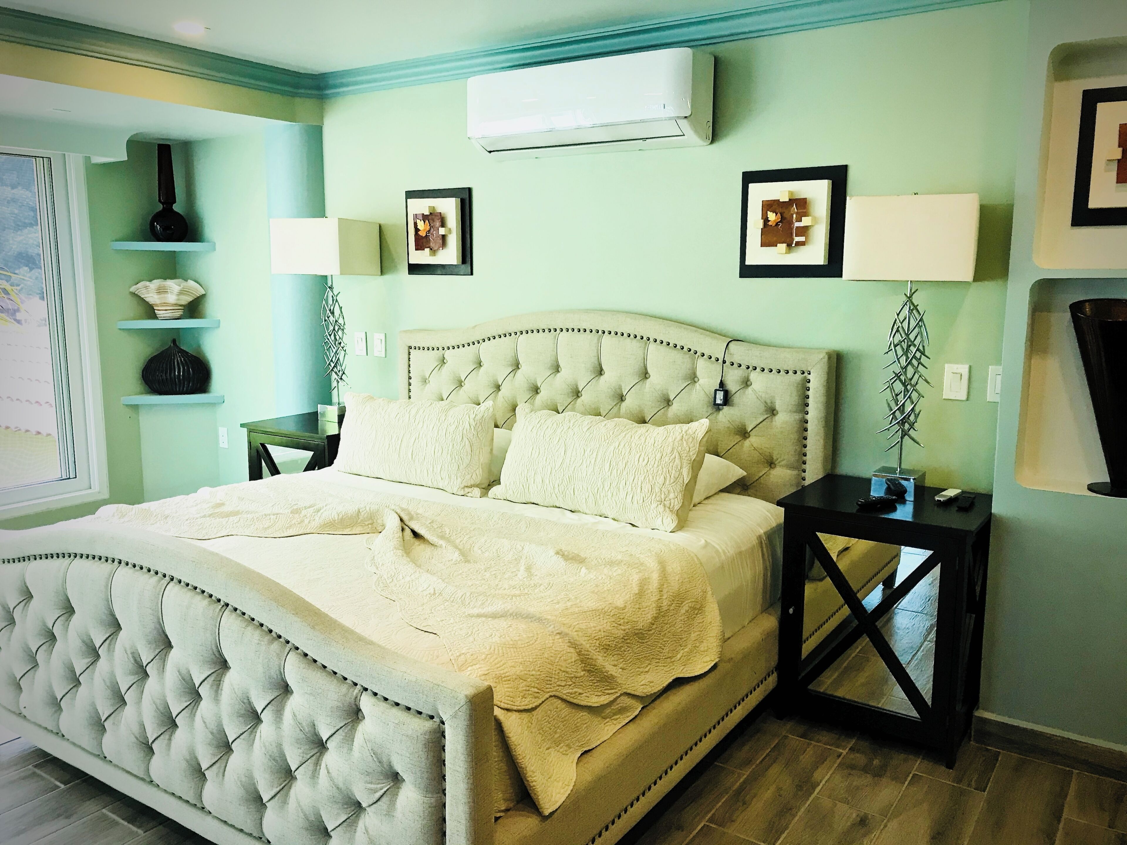 Een slaapkamer met een gestoffeerd hoofdeinde, een bed met een witte dekbedovertrek, twee nachtkastjes met lampen en een airconditioner aan de muur.