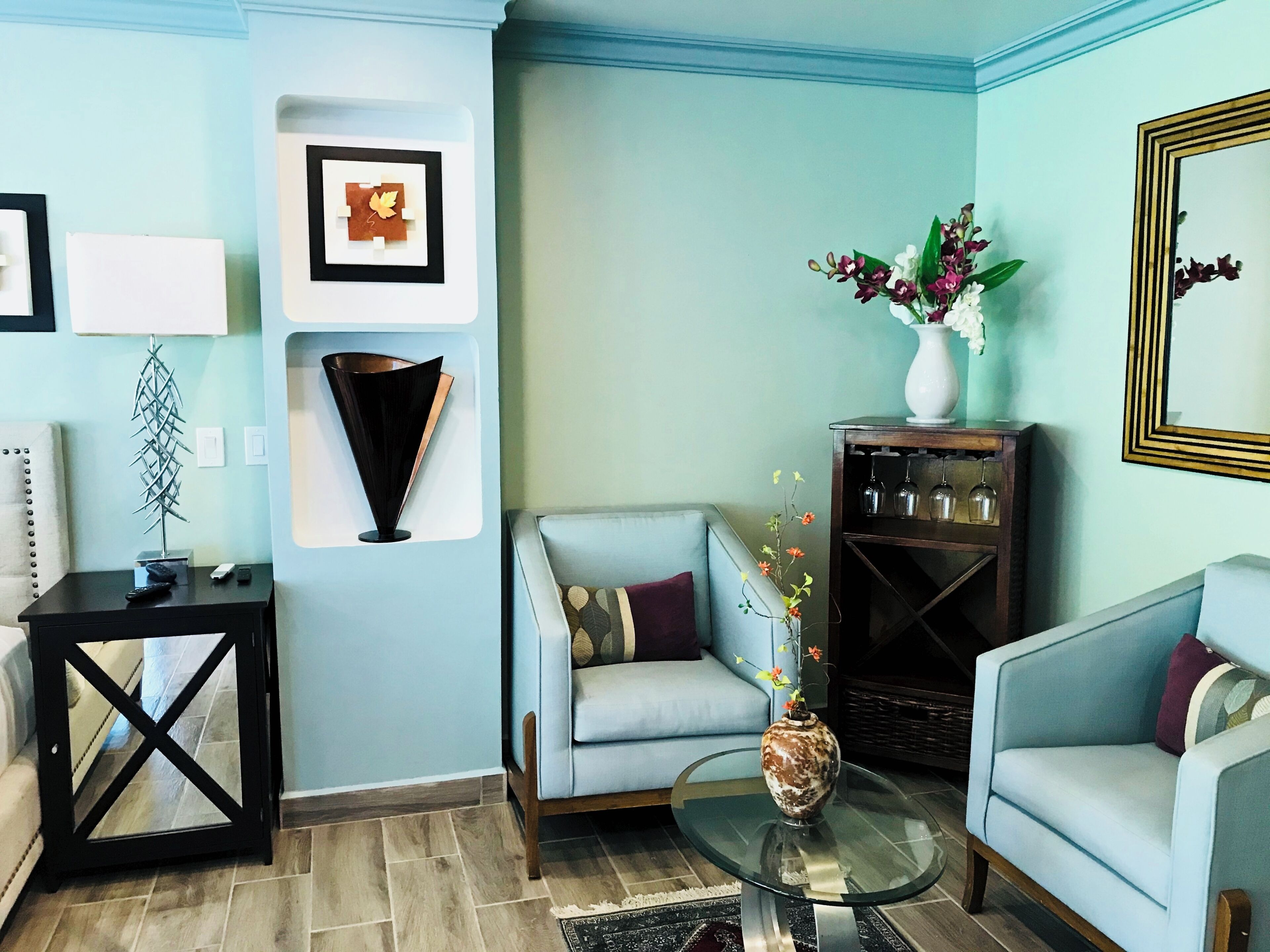 Ein Wohnzimmer mit hellblauen Wänden, einem Glastisch, zwei hellblauen Sofas, einem dunklen Holztisch mit Blumenvase und einem wandmontierten Spiegel.
