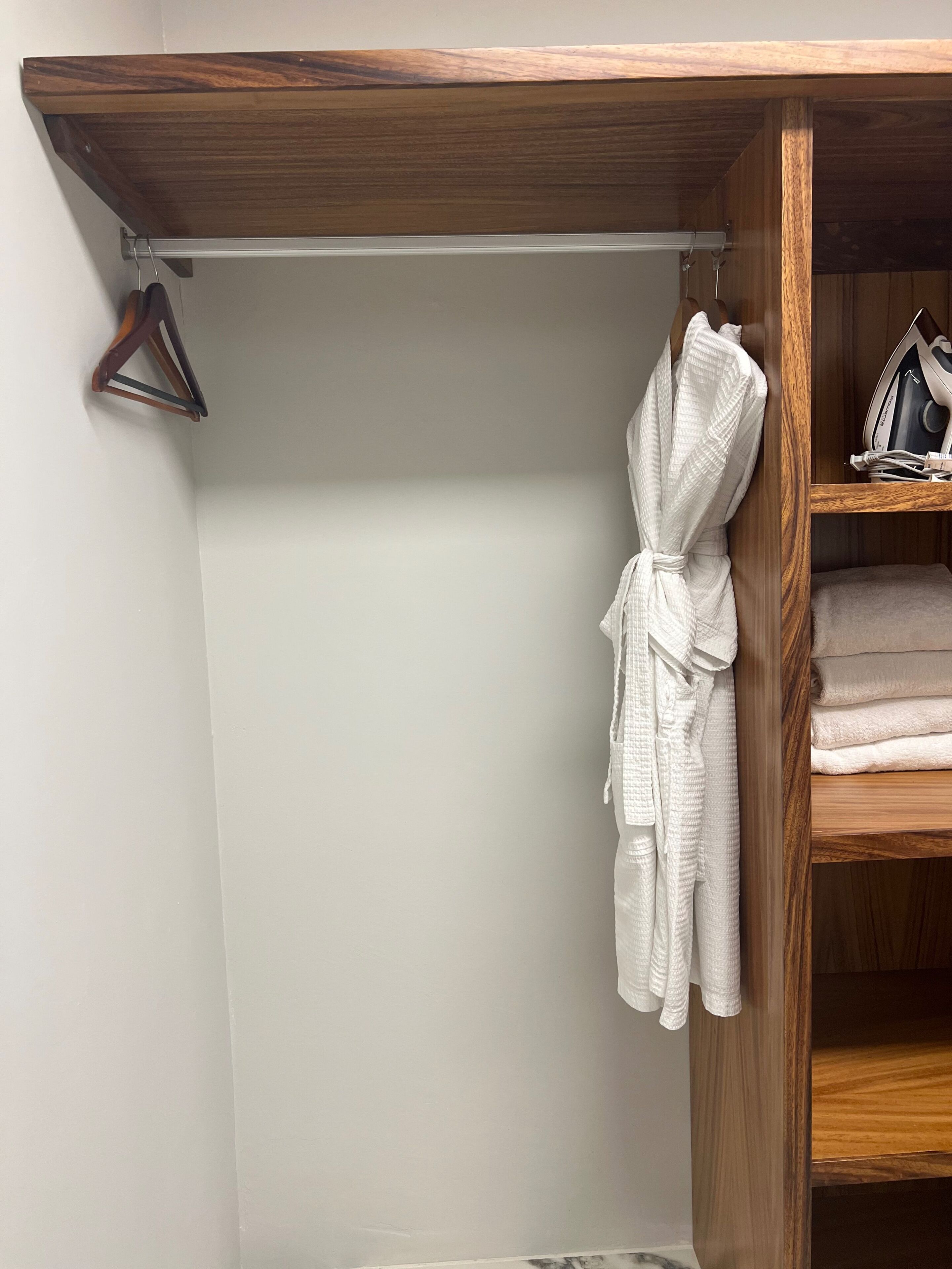 A wooden closet with a robe hanging, a shelf with folded towels, and an iron on the top shelf.