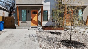 A modern two-story house with a concrete facade, a wooden door, and a small garden area with gravel and a tree.
