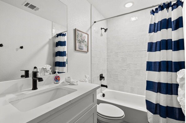 Un baño con lavamanos blanco, grifo negro, inodoro blanco y ducha con cortinas azul y blanco a rayas.