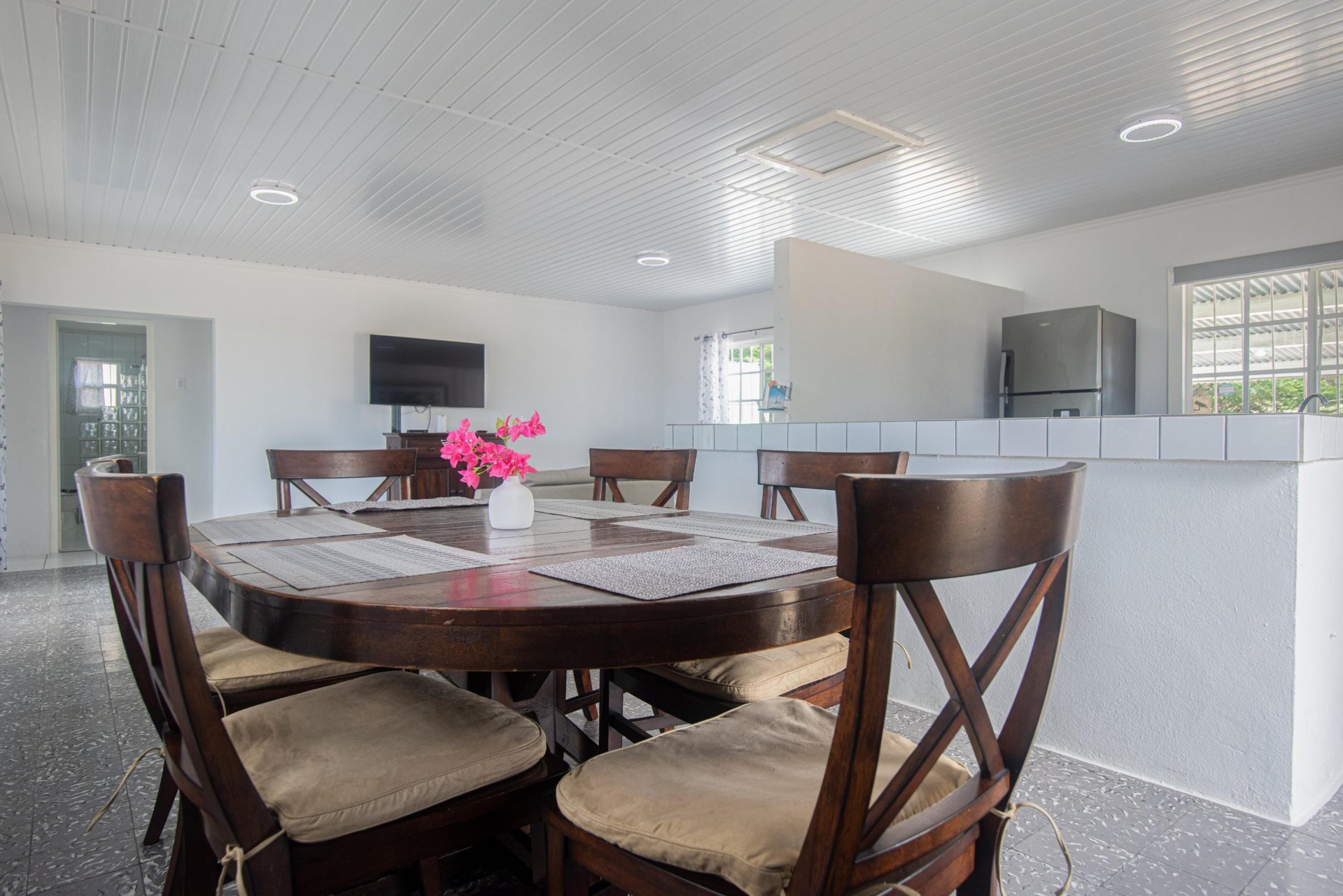 A modern dining area with a round table, six wooden chairs with cushioned seats, a white vase with pink flowers, and a flat-screen TV mounted on the wall.
