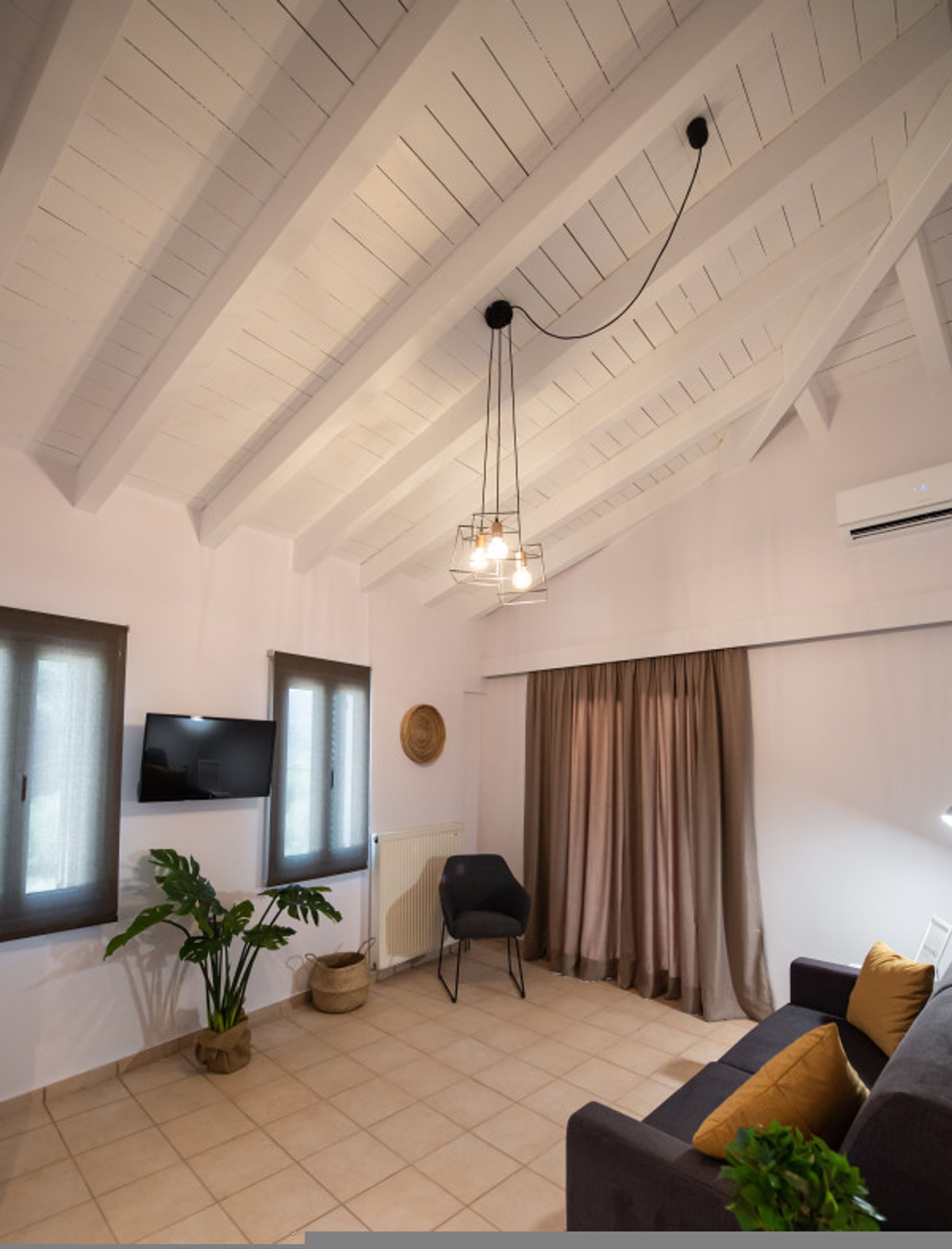 Ein Wohnzimmer mit hoher Decke, einem an der Wand montierten Fernseher, einer Couch mit Kissen, einem Sessel, einer Topfpflanze und einem Fenster mit Vorhängen.