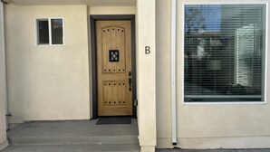 A beige building with a wooden front door, a small window above the door, and a large window with blinds to the right. - NEW 2BR House IN Glendale Built in 2024 With Garage Private Patio p77 (Glendale)