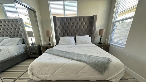 A large bed with a tufted headboard, a bedside table with a lamp, and a window with blinds. - NEW 2BR House IN Glendale Built in 2024 With Garage Private Patio p77 (Glendale)