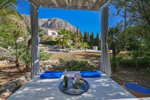 A table set with a bottle, glasses, and flowers, overlooking a mountainous landscape with a pool and greenery.