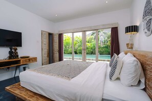 A bedroom with a large bed, a TV, a wooden desk, and a view of a pool and greenery. - Villa Casa del Sol (Canggu)
