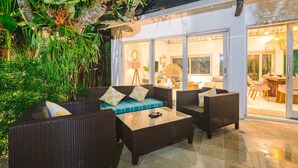 Un patio al aire libre con mobiliario de mimbre, una mesa de centro de vidrio y vista al interior a través de puertas de cristal.