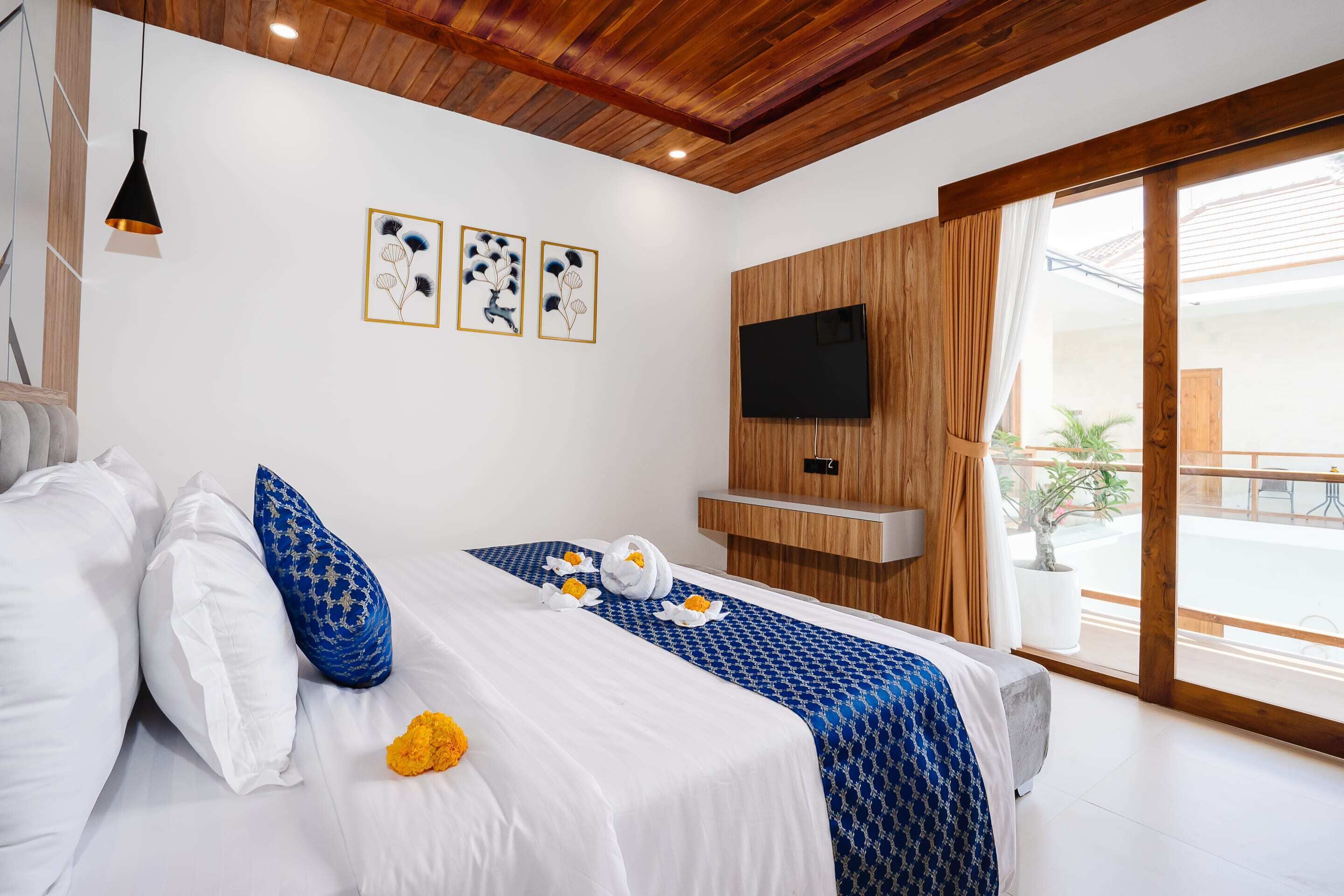 Een moderne hotelkamer met een houten plafond, een groot bed met wit en blauw beddengoed, een flatscreen-tv bevestigd aan een houten paneel, en een balkon met een bloempot.