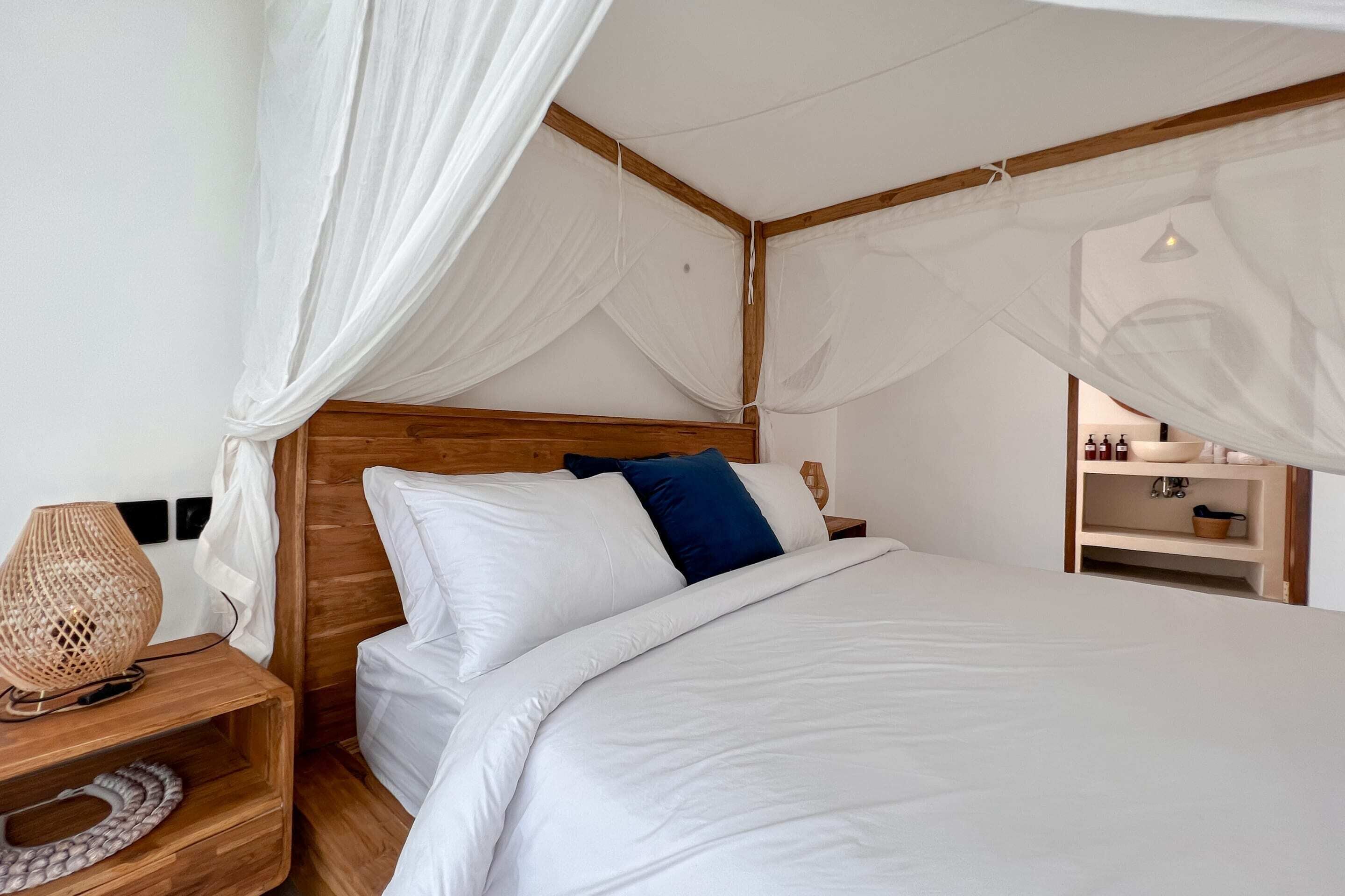 Un dormitorio con cabecera de madera, ropa de cama blanca, almohada azul y una lámpara de noche.