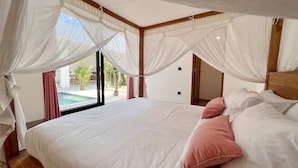 A bedroom with a large bed, white bedding, and a view of a swimming pool. - Villa Timatou (Canggu)
