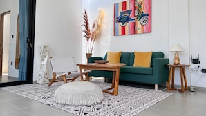 A living room with a green sofa, a wooden coffee table, a white chair, a patterned rug, and a painting of a scooter. - Villa Timatou (Canggu)