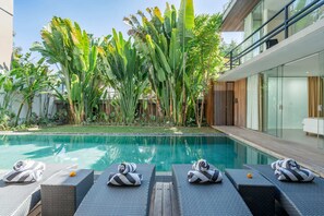 Piscina com espreguiçadeiras e toalhas, rodeada por plantas tropicais e um edifício moderno.