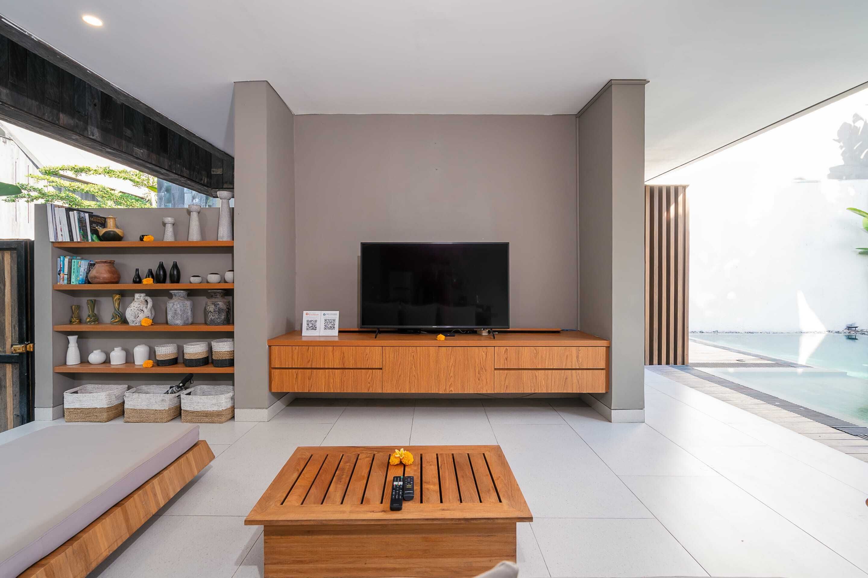 A modern living room with a flat-screen TV on a wooden console, a coffee table with remote controls, and a bookshelf with decorative items.