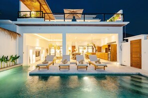A modern house with a swimming pool, lounge chairs, and a well-lit interior. - Villa Mayasa (Munggu)