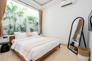 A modern bedroom with a bed, a round black side table, a large mirror, and a window with curtains. - Villa Molly (Canggu)