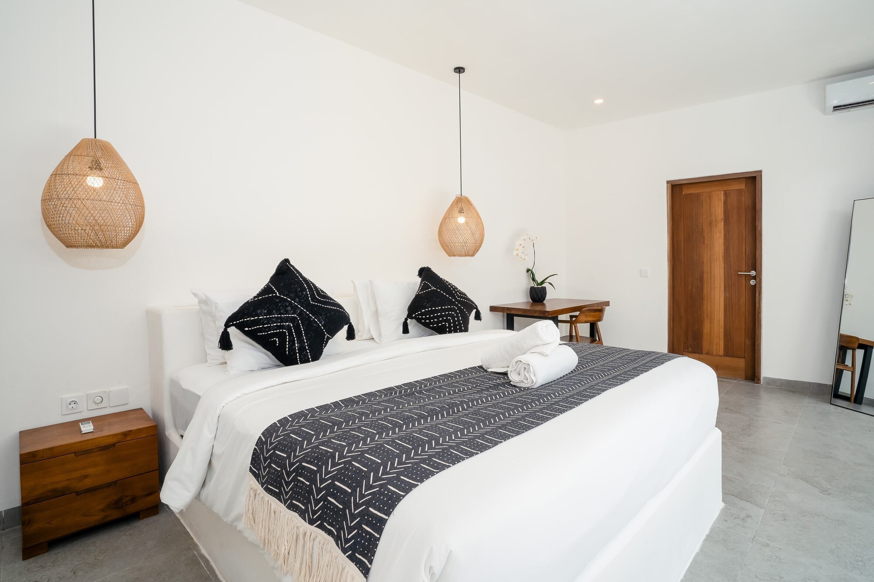 A neatly made bed with a patterned bedspread, two hanging woven light fixtures, a wooden nightstand, a small wooden table, and a wooden door.