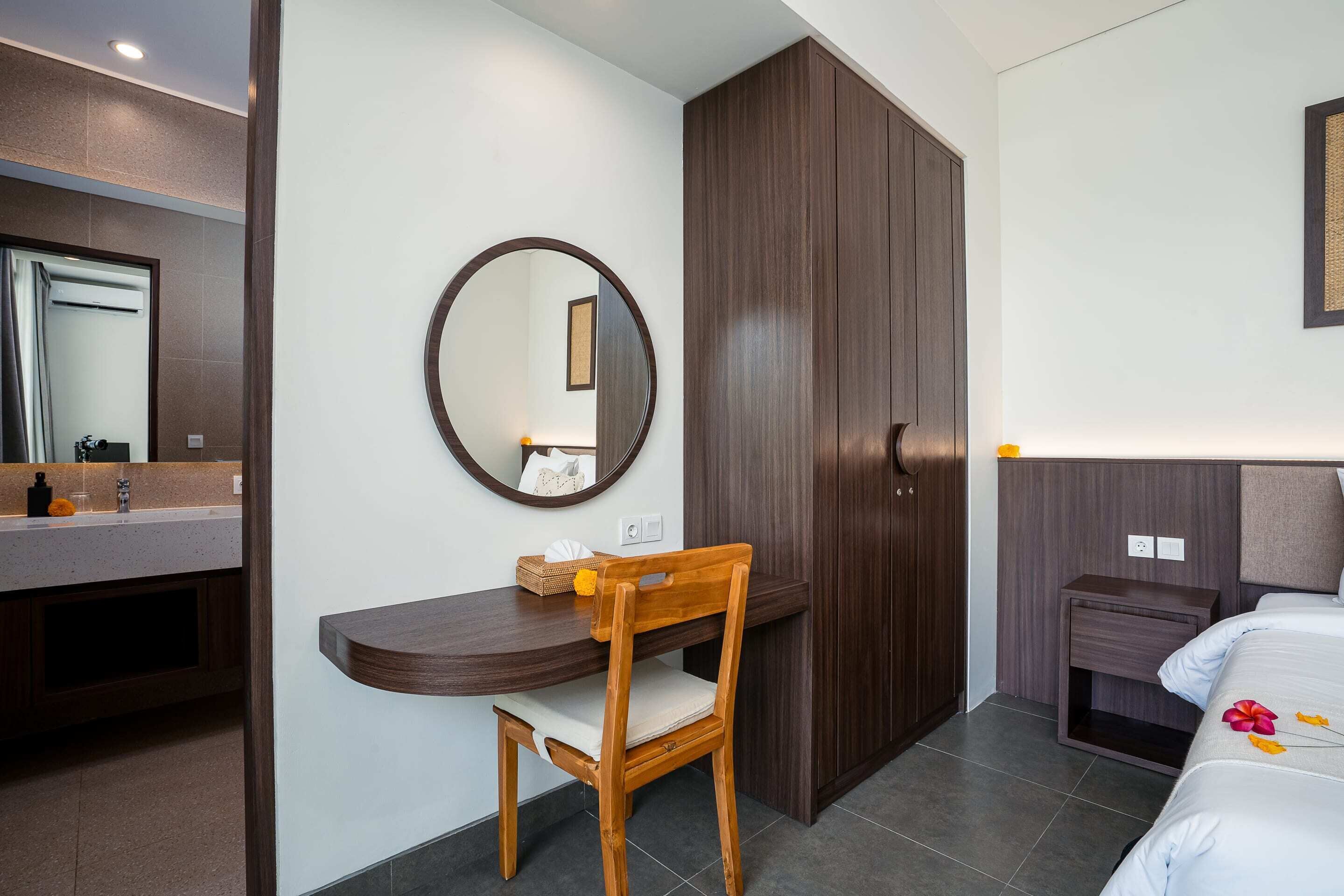 A hotel room with a wooden desk, a chair, a large mirror, a wardrobe, a bedside table, and a bed with a floral decoration.