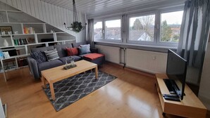 A modern living room with a sofa, coffee table, and TV stand. There is a bookshelf and a window with a view of the city. - Gemütliche Dachgeschosswohnung bei Tutzing am Starnberger See (Tutzing)