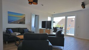 Un salon moderne avec un canapé, une table basse et un téléviseur à écran plat. Une grande fenêtre offre une vue sur l’extérieur.