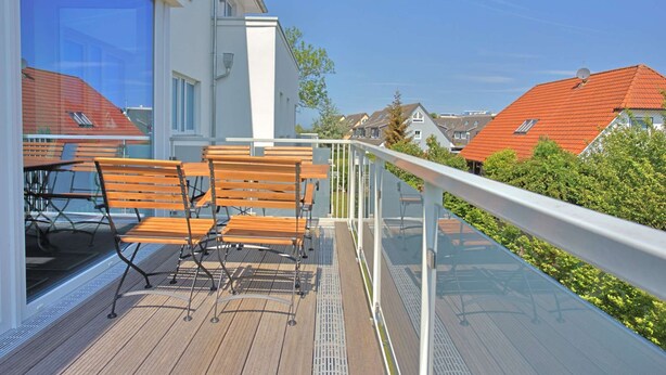 Un balcon doté de meubles en bois, d’une rambarde en verre et offrant une vue sur des maisons aux toits rouges.