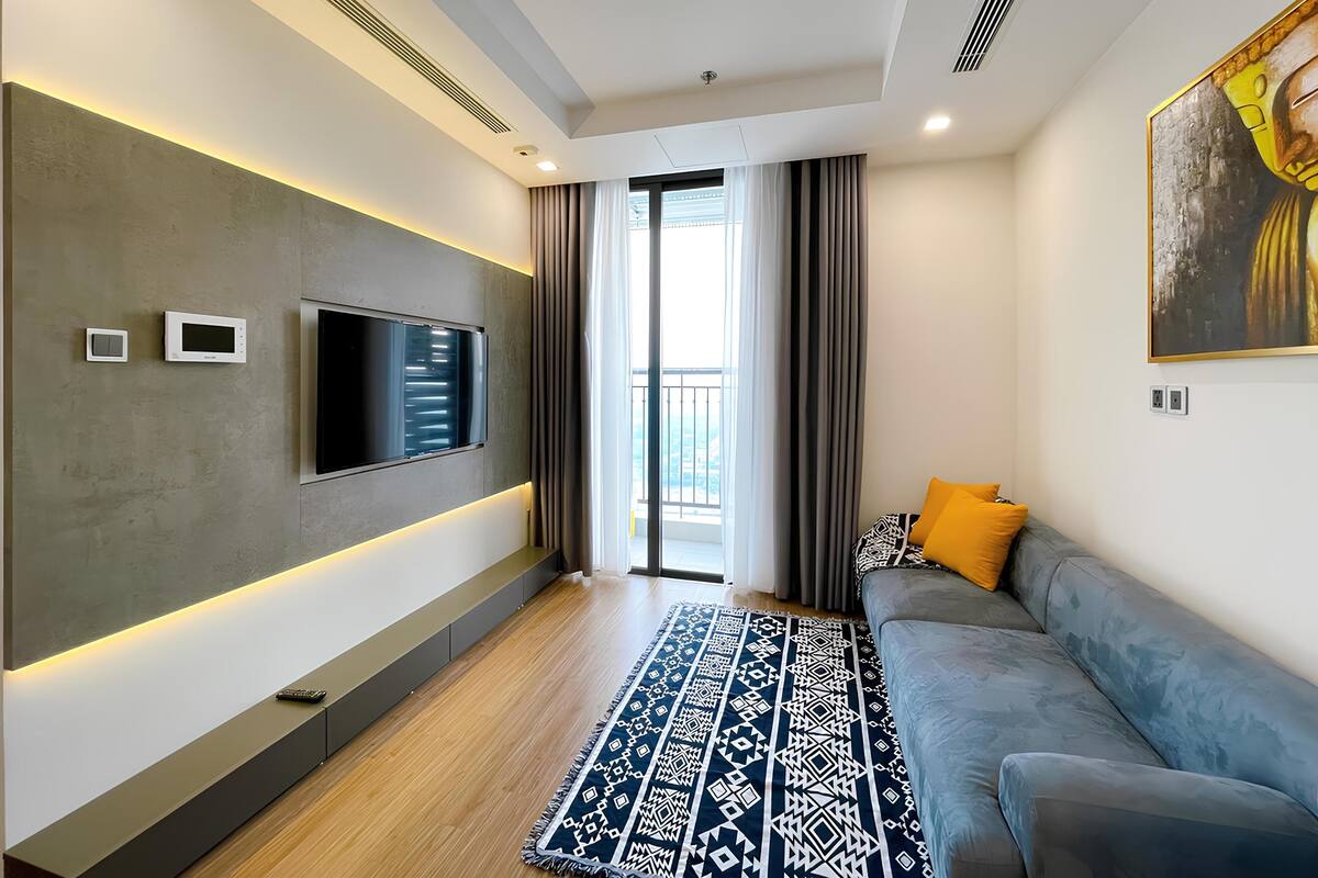 A modern living room with a flat-screen TV mounted on the wall, a grey sofa with orange cushions, a patterned rug, and a large window with curtains.